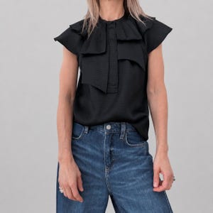 May include: A black short-sleeved blouse with a textured fabric. The blouse features a high, ruffled collar and a layered design on the front. The model is wearing blue denim jeans.