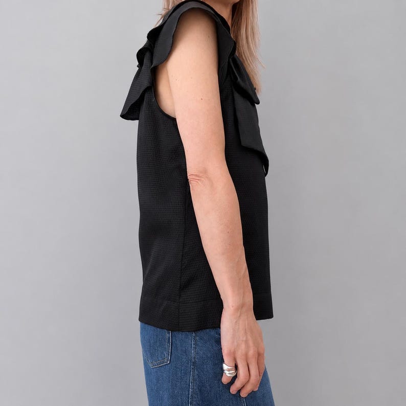 May include: Black sleeveless top with ruffled shoulder details. The top has a textured fabric and a simple, elegant design. The model is wearing blue jeans. The top is a versatile piece for various occasions.