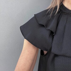 May include: Black top with ruffled sleeves. The top has a textured fabric and a high neckline. The sleeves are short and flared, with a layered ruffle detail. The background is a solid gray.