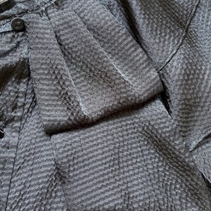 May include: Close-up of a dark gray fabric with a textured, striped pattern. The fabric appears to be part of a garment, possibly a jacket or blouse, with visible seams and a button. The material has a subtle sheen.