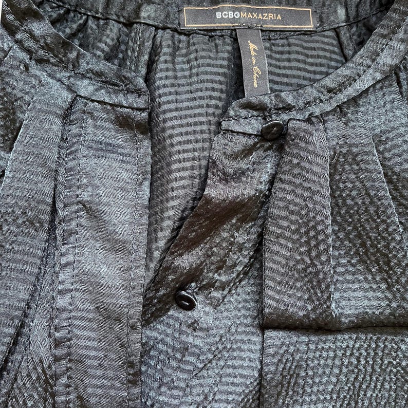 May include: Close-up of a black blouse with a textured pattern and a mandarin collar. The blouse has a button closure and a label that reads "BCBGMAXAZRIA". The fabric has a subtle sheen.