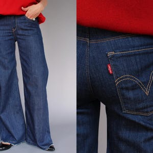 Può includere: Jeans in denim blu scuro con un design a gamba larga. I jeans presentano un classico stile a cinque tasche, con un'etichetta rossa Levi's sulla tasca posteriore. Le cuciture sono di colore beige a contrasto.