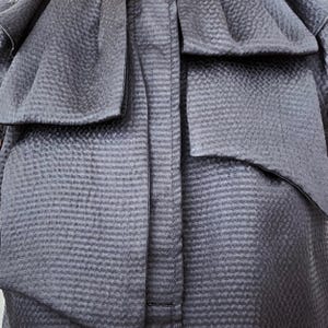 May include: Close-up of a dark gray garment with a textured, woven pattern. The fabric is gathered and folded, creating a layered effect with a prominent bow detail. The stitching is visible along the center.