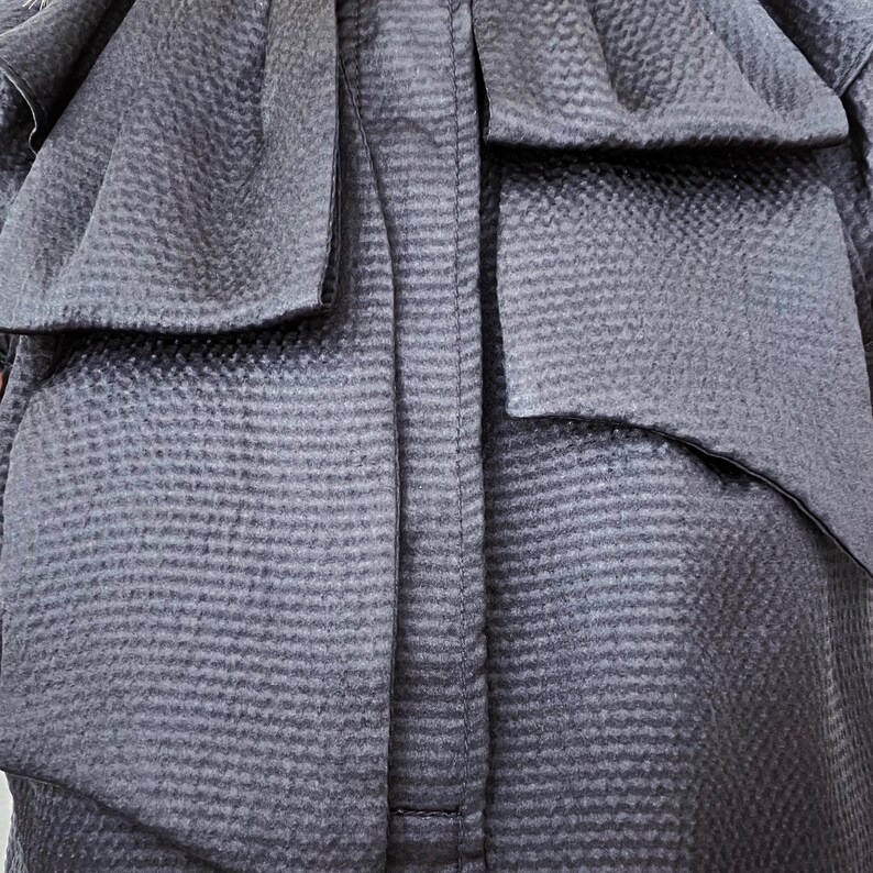 May include: Close-up of a dark gray garment with a textured, woven pattern. The fabric is gathered and folded, creating a layered effect with a prominent bow detail. The stitching is visible along the center.