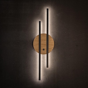 Op de afbeelding: Moderne wandlamp met een cirkelvormige houten basis en zwarte verticale balken. Het licht geeft een zachte gloed, waardoor een minimalistische en stijlvolle verlichtingsarmatuur ontstaat. Geschikt voor moderne woondecoratie. De diameter is ongeveer 15 cm.