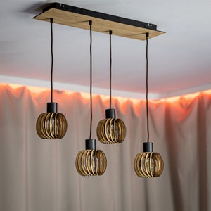 Könnte beinhalten: Eine moderne Pendelleuchte mit vier Lichtern. Die Leuchte hat eine rechteckige Holzbasis mit vier schwarzen Kabeln, an denen kugelförmige Lampenschirme aus Holz hängen. Die Schirme haben ein geschichtetes, offenes Design, das die Glühbirnen freilegt. Die Deckenleuchte ist warm und neutral.