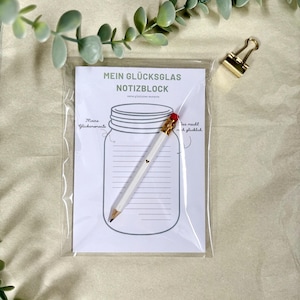 Könnte beinhalten: Ein Notizblock mit dem Text "MEIN GLÜCKSGLAS NOTIZBLOCK" und einer Gläser-Illustration, zusammen mit einem weißen Bleistift mit rotem Radiergummi. Der Notizblock befindet sich in einer transparenten Plastikhülle, mit einer goldenen Büroklammer.