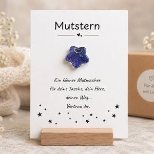 Könnte beinhalten: Eine Grußkarte mit dem Wort "Mutstern" oben, mit einem blauen sternförmigen Anhänger. Die Karte wird von einem Holzständer gehalten. Der Text auf der Karte lautet "Ein kleiner Mutmacher...Vertrau dir." Eine kleine Geschenkbox ist im Hintergrund sichtbar.