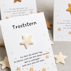 Könnte beinhalten: Weiße Karten mit dem Wort "Troststern" und einer sternförmigen Verzierung. Die Karten haben ein Stern-Design mit kleinen, hellbraunen Sternen, die über die Ränder verteilt sind. Der Text auf der Karte lautet "Ein Stern für dunkle Tage."