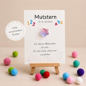 Könnte beinhalten: Eine weiße Karte mit dem deutschen Wort "Mutstern" und Text, präsentiert auf einer kleinen Holzstaffelei. Ein farbenfrohes sternförmiges Ornament ist an der Karte befestigt. Bunte Filzkugeln umgeben die Karte.