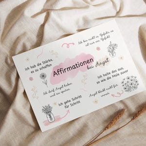 Könnte beinhalten: Eine weiße Karte mit dem deutschen Text "Affirmationen bei Angst" in Rosa und Schwarz, umgeben von floralen Illustrationen und Affirmationen. Die Karte liegt auf einem beigen Stoff.