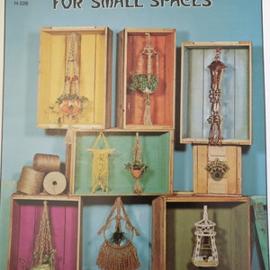 May include: A collection of seven macrame plant hangers displayed in wooden crates. The hangers are made with natural twine and feature different designs, including a bell-shaped hanger, a circular hanger, and a rectangular hanger. The crates are painted in various colours, including yellow, orange, blue, and green. The text "MACRAME' HANGERS FOR SMALL SPACES" is printed at the top of the image. The text "H-228" is printed in the top left corner. The text "30 13117" is printed in the bottom right corner.