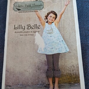 May include: A sewing pattern for a reversible pinafore and leggings for children, featuring a girl wearing the finished garment. The pattern is called "Lilly Belle" and is from Violette Field Threads. The pattern is available in sizes 12 months to 10 years.
