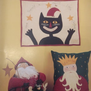 May include: A book titled "Making Spirits Bright" by Cathy Pendleton, featuring illustrations of a black cat wearing a Santa hat, a Santa Claus doll, and a pillow with a king of the elves design.