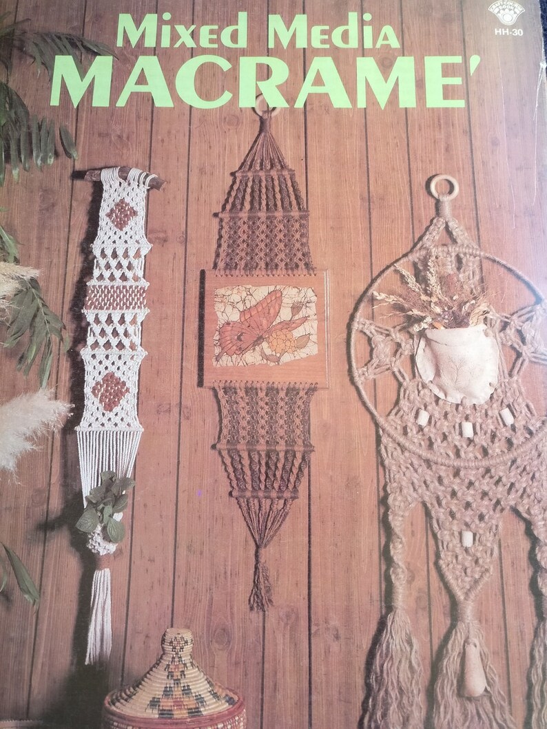 May include: A book cover with the title "Mixed Media Macrame'" in green letters. The cover features three macrame wall hangings, one with a butterfly design, one with a round shape, and one with a rectangular shape. The hangings are made with natural fibers and have a rustic, bohemian aesthetic.