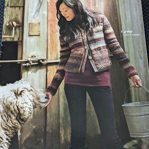 Peut inclure: Une femme portant un cardigan rayé multicolore avec une chemise bordeaux et un jean noir. Elle tient un seau en métal et regarde un mouton blanc. Le texte sur l'image est "Ulla Cupit Geode™ & Jasper™ #276 1 g'day knit in Geode™ BERROCO"