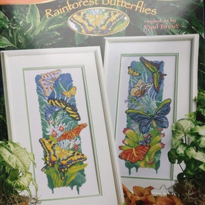 May include: Two framed cross-stitch embroidery designs featuring colorful butterflies in a rainforest setting. The designs are titled "Rainforest Butterflies" and are by artist Paul Brent. The frame size is 12 inches and the design size is 6.75 inches by 16.5 inches.