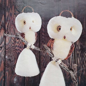 May include: A craft book cover titled Macramé Showcase Classic Charm In Easy-To-Do Designs featuring two fluffy white owls with brown wings perched on a textured branch. The owls have large orange eyes, small brown beaks, and white tails.