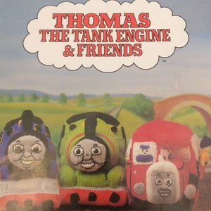 May include: A book cover featuring the title "Thomas the Tank Engine & Friends" in red letters. The cover shows three knitted toy trains: a blue train, a green train, and a red train. The trains have smiling faces and are arranged in a row.