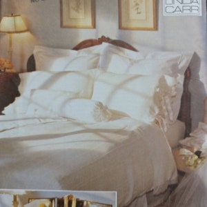 May include: A white bedding set with a ruffled pillow and a white bed skirt. The bedding is on a bed with a wooden headboard. The image also shows a small table with a mirror and a few decorative items.