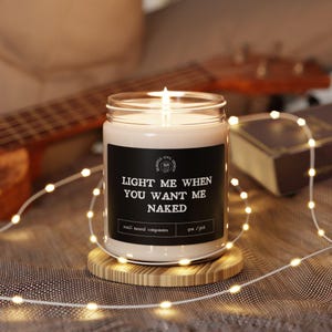 Könnte beinhalten: Eine brennende Kerze in einem Glas mit einem schwarzen Etikett mit der Aufschrift "LIGHT ME WHEN YOU WANT ME NAKED". Die Kerze steht auf einem runden Untersetzer aus Holz. Eine Lichterkette und eine Gitarre sind im Hintergrund.