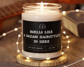 May include: A lit candle in a glass jar with a black label that reads "SMELLS LIKE A BADASS HAIRSTYLIST IN HERE." The candle sits on a wooden coaster and is surrounded by string lights. The candle is 9 oz.