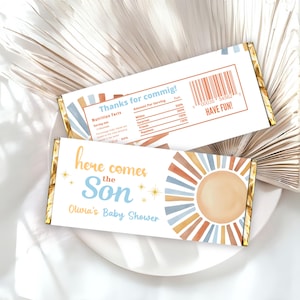 May include: Two chocolate bar wrappers with gold foil edges. One wrapper says "Thanks for commig!" and the other says "Here comes the Son, Olivia's Baby Shower." Both have a sun design in orange, blue, and tan.