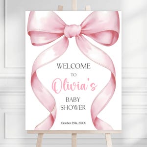 May include: A baby shower welcome sign featuring a large pink bow and flowing ribbons. The sign reads "Welcome to Olivia's Baby Shower" in elegant script and includes the date October 25th, 20XX. The sign is displayed on a wooden easel.