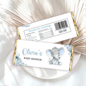 Puede incluir: Dos envoltorios de barra de chocolate con bordes dorados. Un envoltorio tiene el texto "Thanks for commig!" y una tabla de información nutricional. El otro envoltorio dice "Olivia's BABY SHOWER" con una ilustración de elefante en acuarela.