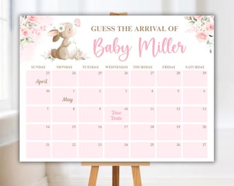 May include: A pink and white baby shower calendar with the text "Guess the Arrival of Baby Miller." The calendar features a cartoon bunny, floral accents, and dates for April and May. It is displayed on a wooden easel.