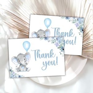 May include: Two white cards with a watercolor illustration of a gray elephant holding a blue balloon. The cards have the text "Thank you!" in light blue script and are decorated with blue floral accents. The cards are on a white plate.