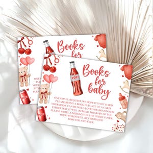 May include: Two baby shower invitation cards with a watercolor design. The cards feature the text "Books for baby" in red script, along with illustrations of teddy bears, soda bottles, and baby clothes. The cards are on a white plate.