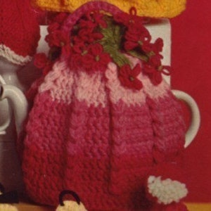 May include: A handmade, knitted tea cozy in shades of pink and red, resembling a handbag. The cozy is adorned with red flower embellishments and a handle. The design is a mix of light and dark pink, creating a gradient effect. The cozy is designed to fit over a teapot.