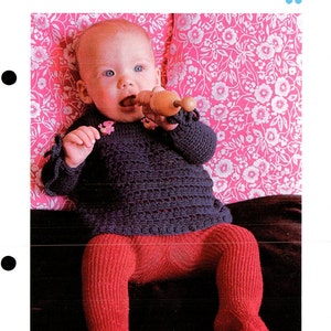 May include: A baby wearing a navy blue openwork top and red tights. The top has long sleeves with decorative ties. The baby is holding a wooden toy. The background is a pink floral pattern.