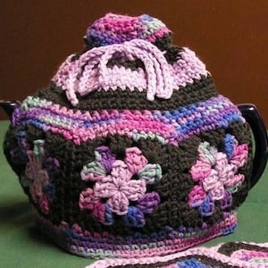 May include: A crocheted teapot cozy in shades of purple, pink, and black. The cozy features a floral design and is made with a variety of stitches.