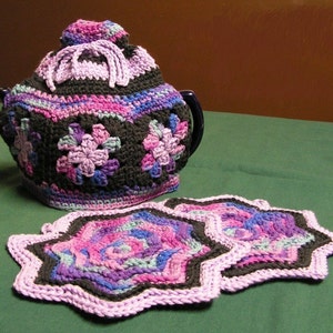 Puede incluir: Un calentador de té de ganchillo y dos posavasos a juego. El calentador de té es negro con detalles florales rosas, morados y azules. Los posavasos tienen forma de estrella con colores similares. El calentador tiene una tapa con asa.