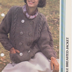 May include: A brown double-breasted knitted jacket with a cable knit pattern and large buttons. The jacket is worn by a person sitting on the ground with a white skirt.