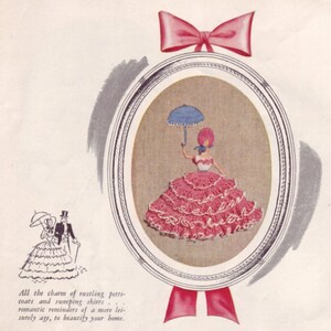 May include: A framed embroidery depicting a woman in a pink dress holding a blue and white umbrella. The embroidery is surrounded by a silver frame and a pink ribbon.