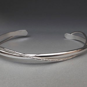 May include: A silver-colored metal cuff bracelet featuring a bypass design where two bands overlap. One band has a smooth, polished finish, while the other band displays a textured, hammered surface. The bracelet has an open back design.