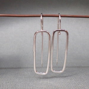 May include: A pair of silver dangle earrings with a rectangular shape. The earrings are made of a thin wire and have a simple, modern design.