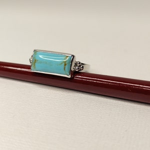 May include: A rectangular turquoise ring with a silver band. The turquoise stone has brown veining. The ring is set on a dark red surface. The ring is a piece of jewelry.
