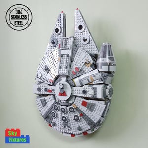 May include: A white and grey LEGO Millennium Falcon model, with red accents, mounted on a wall. The model is made of plastic and has a detailed design. The text "304 STAINLESS STEEL" is visible in a circle on the wall.
