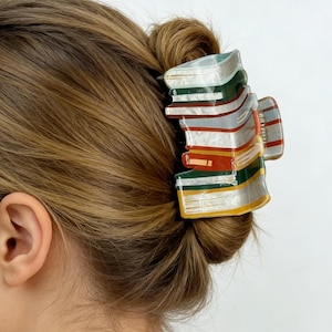 May include: A colorful hair claw clip in the shape of stacked books. The clip features a variety of colors, including green, red, yellow, and white, and is attached to a bun hairstyle. The clip is made of a shiny material.