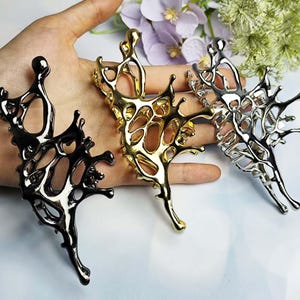 May include: Three abstract metal hair clips in black, gold, and silver. Each clip features an open, organic design with a flowing, asymmetrical shape. The clips are displayed on a hand, showcasing their form and color variations.