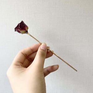 Può includere: Un bocciolo di rosa essiccato attaccato a un bastoncino di metallo dorato.