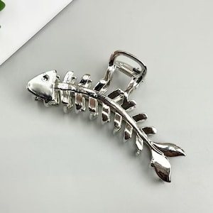 May include: Silver-colored hair claw clip in the shape of a fish skeleton. The clip is made of metal and features a detailed design of a fish's bones, head, and tail. The clip is designed to hold hair in place.