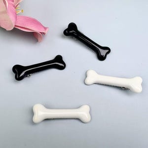 May include: Four bone-shaped hair clips in black and white. The clips are arranged on a light blue surface, with a pink flower in the upper left corner. The clips are made of a glossy material.