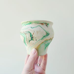 May include: A small ceramic planter featuring a marbled pattern with swirls of cream, light green, and dark green colors. The vessel has a rounded body that tapers toward the base and a wide, smooth rim, showcasing a unique blend of colors.
