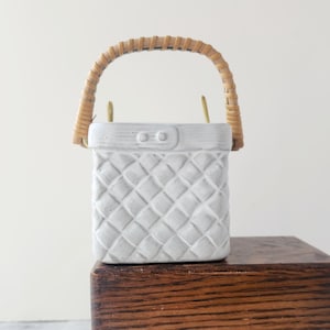 May include: A white ceramic basket with a woven rattan handle. The basket has a quilted pattern and a rectangular shape. The handle is arched and attached to the top of the basket with gold-colored metal pieces.