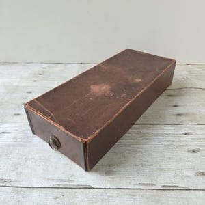 May include: A rectangular brown storage box with a weathered, matte finish and a single circular metal pull handle centered on the front panel. The box features visible signs of wear and texture across its surface, with a simple, functional design.
